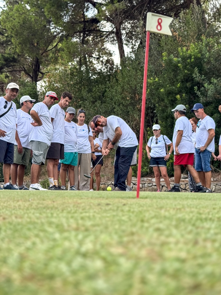 Un partecipante prova un colpo di golf vicino alla bandierina della buca 8, assistito davanti al gruppo del Progetto Icaro, Marelba 2026