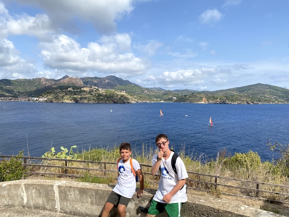 Due partecipanti con magliette Icaro e zaini durante la visita a Forte Focardo, con il golfo e le colline sullo sfondo, Marelba 2026