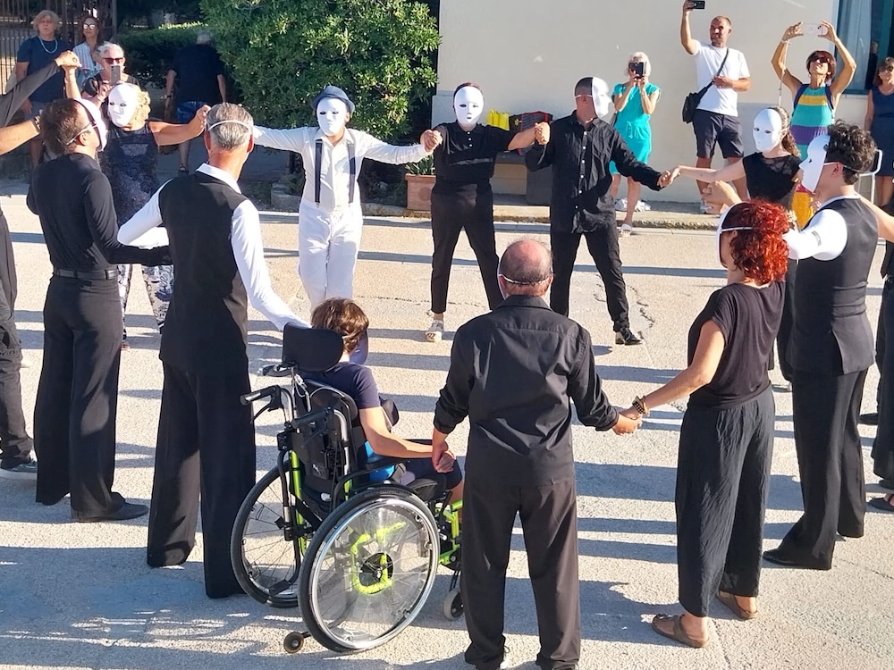 Danzatori, alcuni con maschere bianche, si tengono per mano in cerchio insieme a un partecipante in carrozzina, Marelba 2026