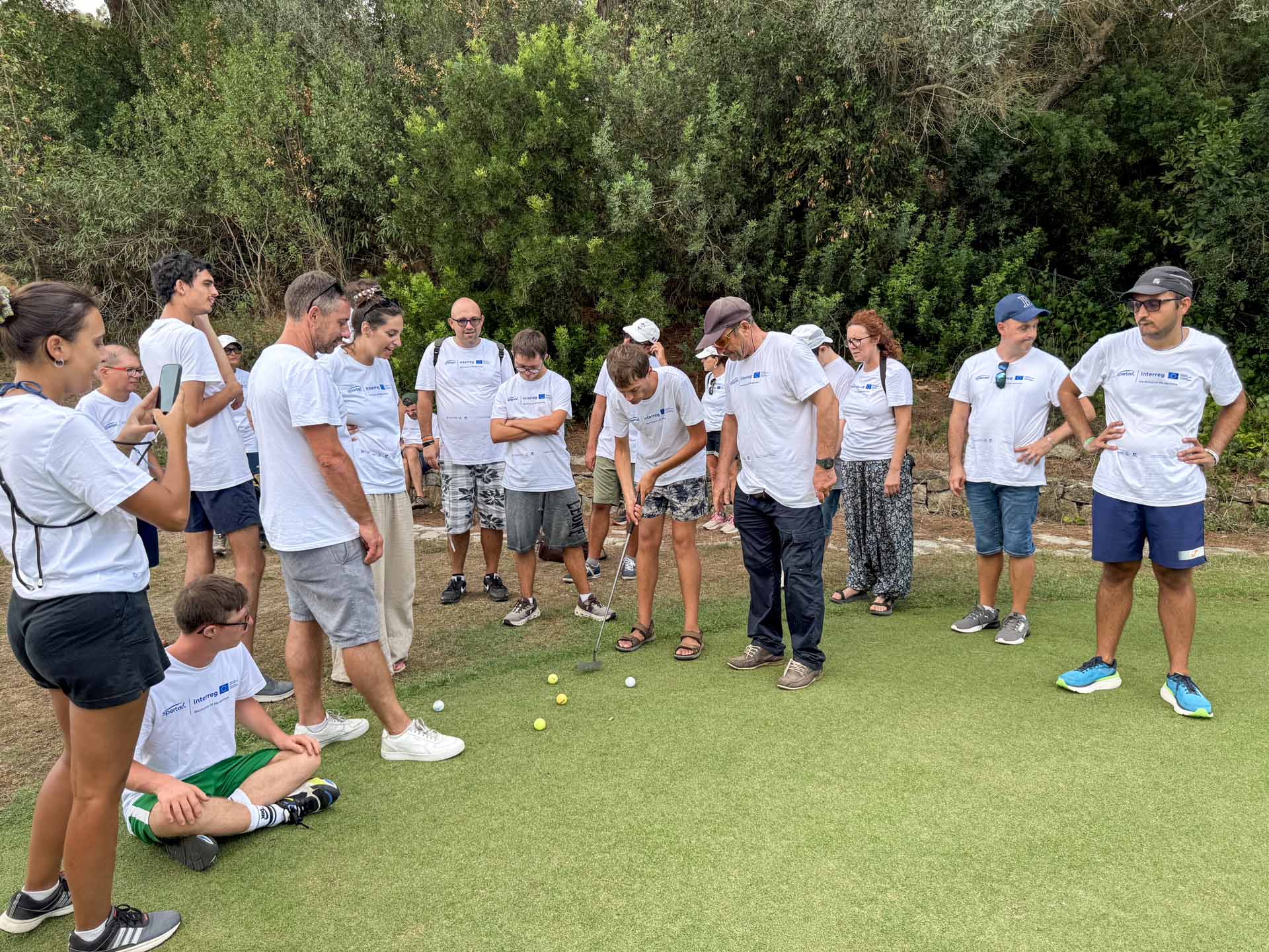 Una partecipante prova il putt con un accompagnatore mentre il gruppo osserva sul green dell’Acquabona, Marelba 2026