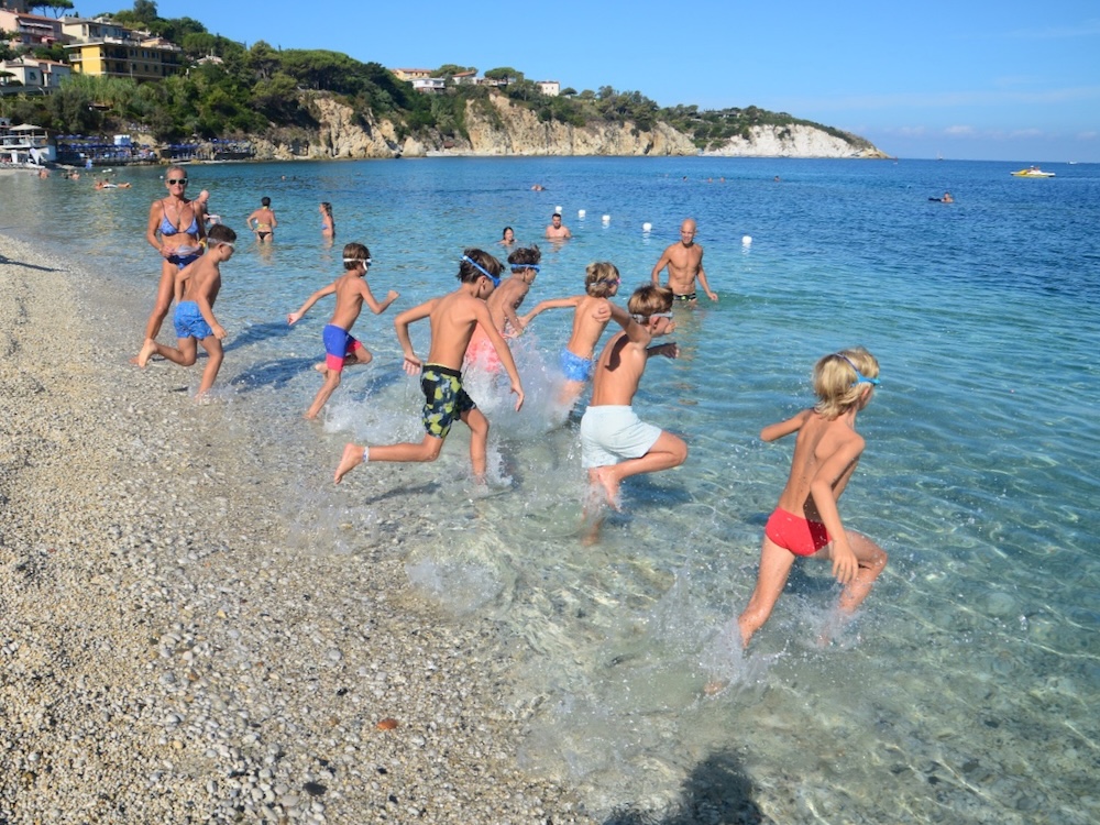 Bambini con gli occhialini corrono dalla spiaggia di ciottoli verso il mare alla partenza della prova di nuoto di Marelba 2026
