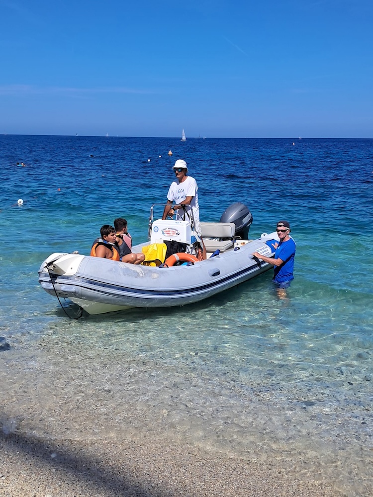 Partecipanti a bordo di un gommone vicino alla spiaggia, con un accompagnatore in acqua, durante le attività di vela di Marelba 2026
